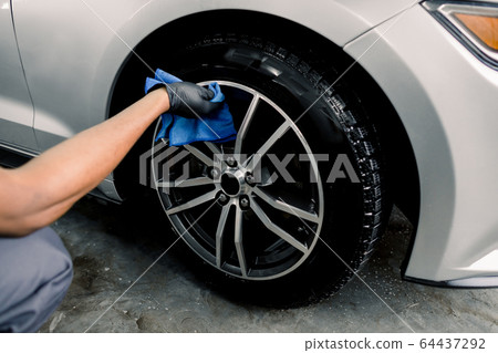 Car rims cleaning, car detailing wash concept. Cropped close up photo of male hand in black rubber glove with blue microfiber cloth washing car alloy wheel at car wash service Car rims cleaning, car detailing wash concept. Cropped close up photo of male hand in black rubber glove with blue microfiber cloth washing car alloy wheel at car wash service 64437292