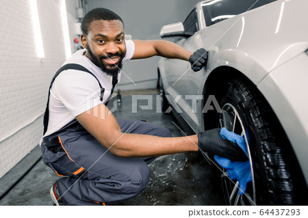 Manual car wash in car wash shop service. Handsome young dark skinned male employee worker cleaning the wheel rim of modern white car by blue microfiber cloth, looking at camera Manual car wash in car wash shop service. Handsome young dark skinned male employee worker cleaning the wheel rim of modern white car by blue microfiber cloth, looking at camera 64437293