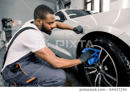 Car washing and detailing photo. African man worker in protective overalls and rubber gloves, washing car alloy wheel on a car wash, using microfiber cloth and special cleaner Car washing and detailing photo. African man worker in protective overalls and rubber gloves, washing car alloy wheel on a car wash, using microfiber cloth and special cleaner 64437294
