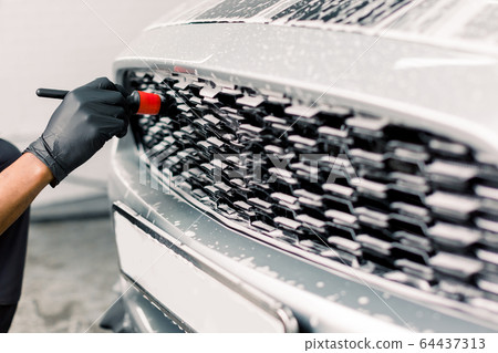 Car detailing wash. Detailing cleaning with soap at car wash. Cropped image of hand of male worker in rubber gloves washing car radiator grille of luxury white car with special brush Car detailing wash. Detailing cleaning with soap at car wash. Cropped image of hand of male worker in rubber gloves washing car radiator grille of luxury white car with special brush 64437313