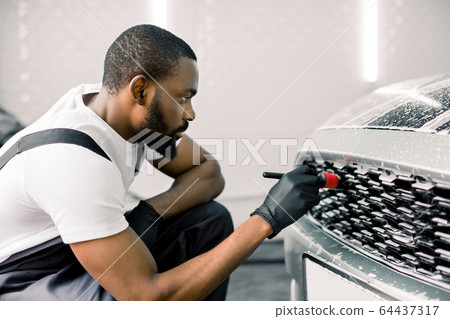 Close up photo of young African male worker in black protective gloves cleaning radiator grille in foam with special brush. Carwash and detailing. Washing machine at the station. Car washing concept. 64437317