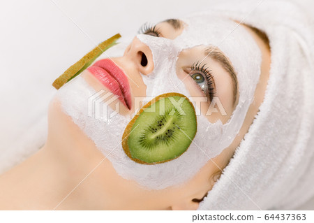 Facial treatment, skin care, home spa. Close up side view photo of young woman in towel on her head, with natural homemade fruit facial mask on skin and slices of fresh kiwi on cheeks. Facial treatment, skin care, home spa. Close up side view photo of young woman in towel on her head, with natural homemade fruit facial mask on skin and slices of fresh kiwi on cheeks. 64437363