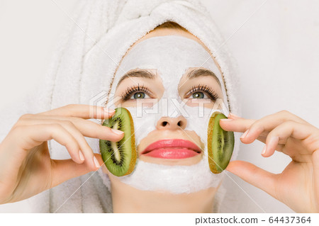 Close up of pretty smiling woman with white moisturizing mask and pieces of fresh kiwi fruit on cheeks. Beauty facial mask, skin care and treatment concept 64437364