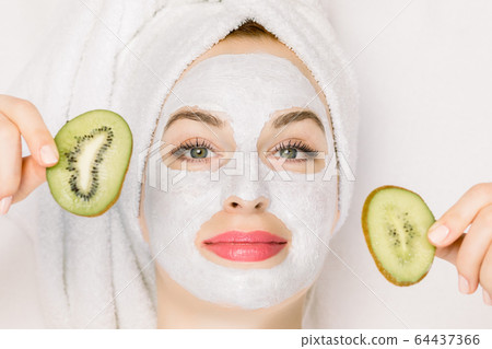 Beautiful woman in bath towel on her head, with purifying white mask on face, lying over white background and holding slices of kiwi fruit. Top view, close up macro photo, copy space 64437366