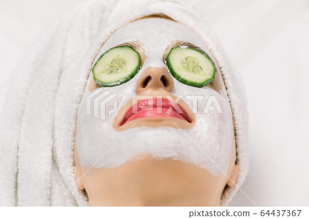 Close up image of young woman with hair wrapped in bath towel, with clay facial mask and cucumber slices on her eyes, lying on isolated white background. Top angle view, macro, copy space Close up image of young woman with hair wrapped in bath towel, with clay facial mask and cucumber slices on her eyes, lying on isolated white background. Top angle view, macro, copy space 64437367
