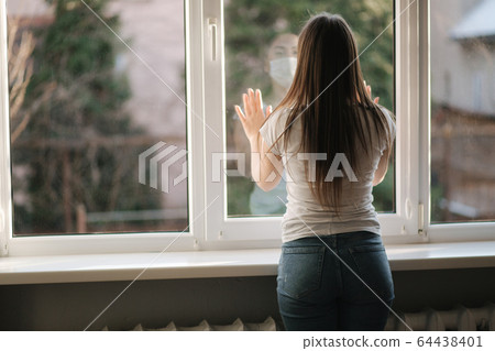 Back view of young woman in medical mask stay by the window in quarantine. Woman looking through the window. Covid-19 Back view of young woman in medical mask stay by the window in quarantine. Woman looking through the window. Covid-19 64438401