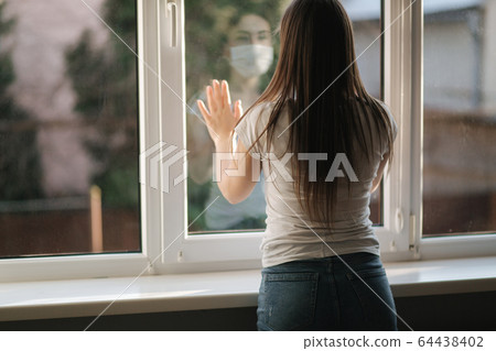 Back view of young woman in medical mask stay by the window in quarantine. Woman looking through the window. Covid-19 Back view of young woman in medical mask stay by the window in quarantine. Woman looking through the window. Covid-19 64438402