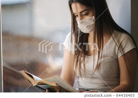 Young woman sits on windowsill in protective mask and look out the window. Alone at home in quarantine. Woman read the book. Coronavirus theme. Stay at home Young woman sits on windowsill in protective mask and look out the window. Alone at home in quarantine. Woman read the book. Coronavirus theme. Stay at home 64438403