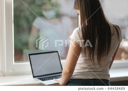 Back view of Female in mask working at home. Woman put laptop on windowsill and work Back view of Female in mask working at home. Woman put laptop on windowsill and work 64438489