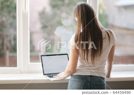 Back view of Female in mask working at home. Woman put laptop on windowsill and work Back view of Female in mask working at home. Woman put laptop on windowsill and work 64438490