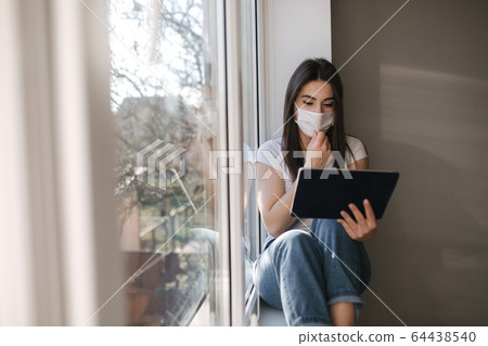 Attractive young woman in white medical mask sits on windowsiil and use tablet. Female have a rest at home in quarantine 64438540