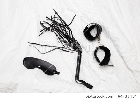 Black sex toys on white sheets of a bed, Leather whip and handcuffs 64439414
