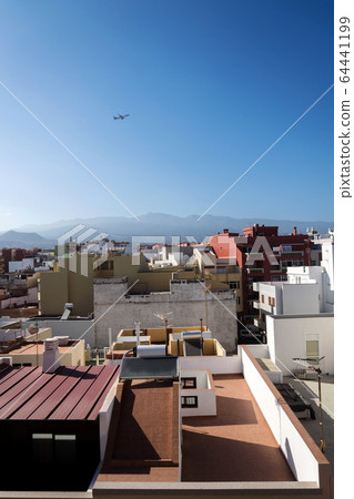 Charter plane leaving Tenerife airport during sunrise seen from roofs of traditional houses with Mount Teide volcano in background, Canary Islands, Spain, sunny blue sky with copy space 64441199