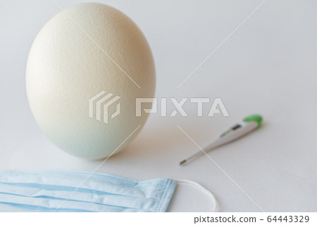 Large white easter ostrich egg, medical mask and thermometer. Egg, respirator and thermometer. Easter and Coronavirus, Covid-19. Ostrich egg, medical mask and thermometer on a white background. 64443329