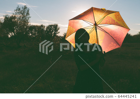 A girl with a colored umbrella poses in a field. The umbrella is illuminated from behind by the sunset light 64445241