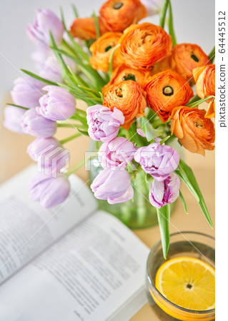 Spring composition with a lilac tulips and orange buttercups and cup of green tea with lemon, an open book on a wooden table. Stay at home concept 64445512