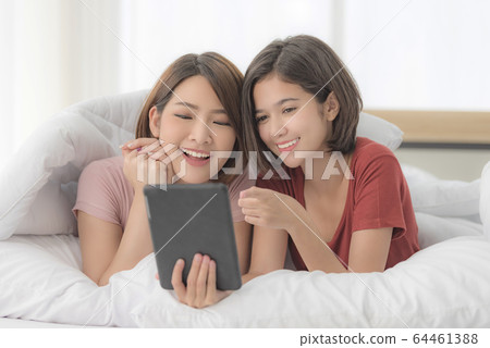 Young asian girl lying on the bed next to her elder sister, both being covered under the blanket, and watching the video on tablet Young asian girl lying on the bed next to her elder sister, both being covered under the blanket, and watching the video on tablet 64461388
