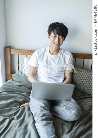 Lifestyle image of a young man in his 20s sitting on a bed and operating a computer Lifestyle image of a young man in his 20s sitting on a bed and operating a computer 64464290