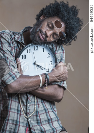 A curly-haired Dominican with open glasses on his glasses embraces a large wall clock and smiles 64469523
