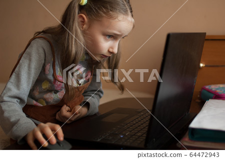 Serious little girl is concentrated in front of her black laptop while focused on doing homework during the quarantine because of the pandemia of Coronavirus Covid-19. Remote education 64472943
