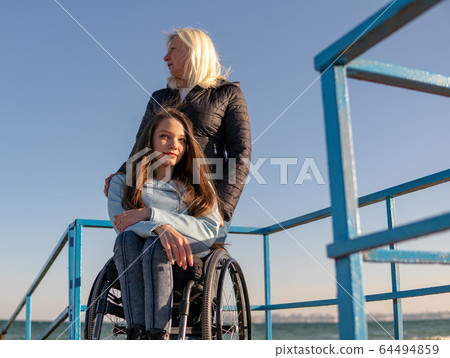 Young disabled woman in wheelchair with her mother walking near the sea on the ramp for people with disabilities. Family leisure Young disabled woman in wheelchair with her mother walking near the sea on the ramp for people with disabilities. Family leisure 64494859