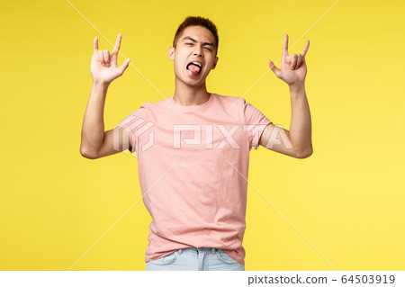 Lifestyle, education and people concept. Enthusiastic handsome and cool asian man in pink t-shirt, show tongue and rock-n-roll sign, having fun, partying, standing yellow background 64503919