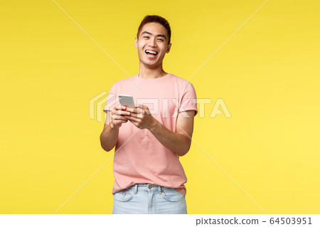 Technology, communication and lifestyle concept. Enthusiastic asian male in pink t-shirt laughing from cool joke or video, using mobile phone, smiling pleased camera, stand yellow background Technology, communication and lifestyle concept. Enthusiastic asian male in pink t-shirt laughing from cool joke or video, using mobile phone, smiling pleased camera, stand yellow background 64503951