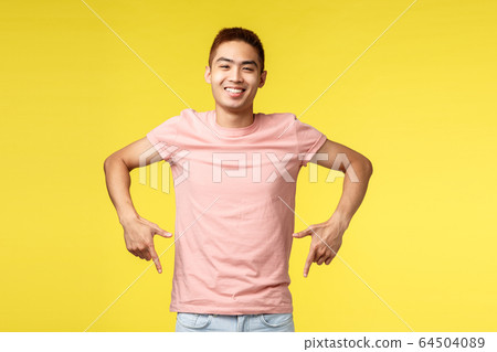 Portrait of easy-going joyful asian guy in pink t-shirt, inviting visit event, pointing fingers down, smiling pleased, recommend product, showing bottom promo, standing yellow background 64504089