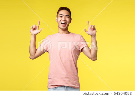 Portrait of young cheerful asian guy in pink t-shirt, pointing fingers up, showing way to product, smiling amused and happy, give advice, recommend item, yellow background 64504098