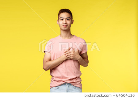 People, different emotions and lifestyle concept. Smiling touched asian man with stylish haircut, pink t-shirt, hold hands on heart and looking camera pleased, going vacation, yellow background People, different emotions and lifestyle concept. Smiling touched asian man with stylish haircut, pink t-shirt, hold hands on heart and looking camera pleased, going vacation, yellow background 64504136