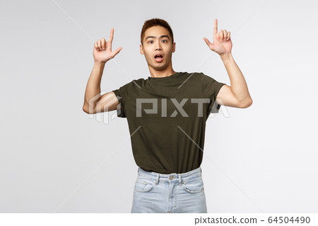 Portrait of shocked and nervous asian man look at camera surprised, pointing fingers up, showing something strange and concerning, asking question about item, grey background 64504490
