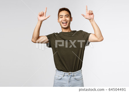 Portrait of optimistic, happy asian man in green t-shirt inviting people check-out advertisement, laughing and smiling excited, pointing fingers up, showing way to banner, grey background Portrait of optimistic, happy asian man in green t-shirt inviting people check-out advertisement, laughing and smiling excited, pointing fingers up, showing way to banner, grey background 64504491