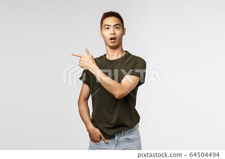Portrait of surprised, curious asian male staring camera startled, asking question and pointing finger left at interesting strange item in store, showing way, standing grey background 64504494