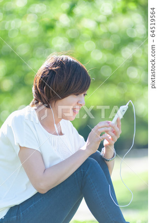 A woman operating a smartphone while sitting in the shade of a tree and listening to music 64519564