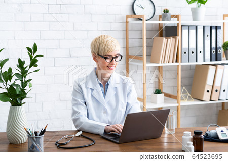 Pretty woman doctor in office at table with laptop Pretty woman doctor in office at table with laptop 64523695