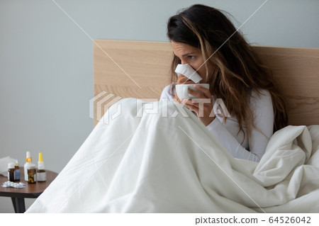 Sick young woman blowing nose, holding handkerchief, sitting in bed Sick young woman blowing nose, holding handkerchief, sitting in bed 64526042