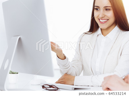 Beautiful young woman sitting at the desk with computer in sunny office. Looks like student girl or business lady communicating with casual dressed man Beautiful young woman sitting at the desk with computer in sunny office. Looks like student girl or business lady communicating with casual dressed man 64533214