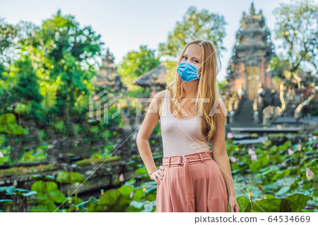 Young woman in medical mask traveler in the background of Pura Taman Kemuda Saraswati Temple in Ubud Young woman in medical mask traveler in the background of Pura Taman Kemuda Saraswati Temple in Ubud 64534669