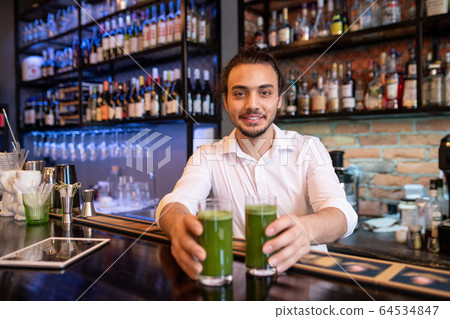 Happy young waiter or barman in white shirt passing you two glasses of smoothie Happy young waiter or barman in white shirt passing you two glasses of smoothie 64534847