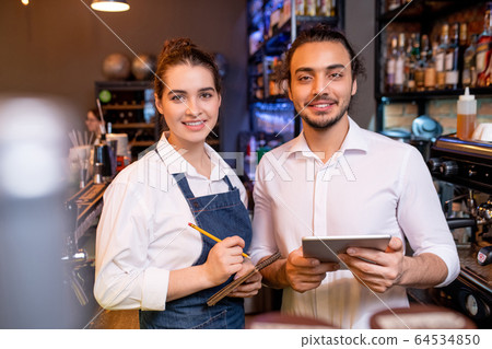 Two young cheerful workers of classy restaurant or cafeteria meeting guests Two young cheerful workers of classy restaurant or cafeteria meeting guests 64534850