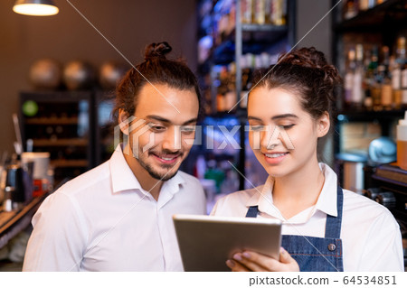 Happy young waitress with tablet showing her colleague new points in online menu 64534851