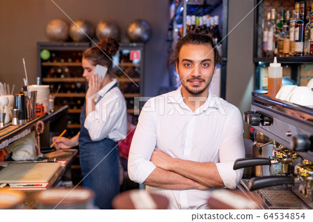 Young handsome waiter in white shirt standing by coffee machine in cafeteria Young handsome waiter in white shirt standing by coffee machine in cafeteria 64534854