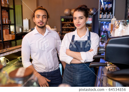Two young colleagues in white shirts and denim aprons standing by workplace 64534856