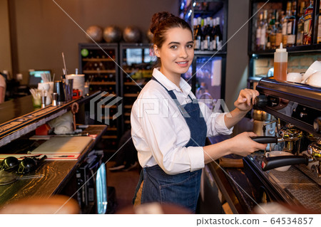 Young friendly waitress in workwear preparing cappuccino by coffee machine 64534857