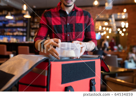 Young courier putting plastic container with cooked food into big open bag 64534859