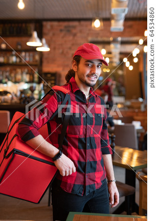 Young courier in red cap and flannel carrying big bag on shoulder Young courier in red cap and flannel carrying big bag on shoulder 64534860