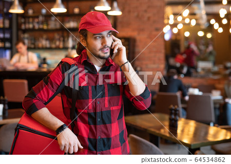 Busy courier in workwear talking to one of clients on mobile phone Busy courier in workwear talking to one of clients on mobile phone 64534862