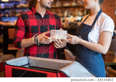 Young courier in red flannel taking container with resh food ordered by client Young courier in red flannel taking container with resh food ordered by client 64534863