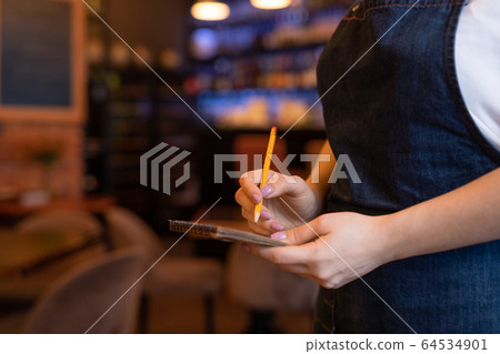 Hands of young waitress in dark blue apron holding pencil over page of notepad Hands of young waitress in dark blue apron holding pencil over page of notepad 64534901