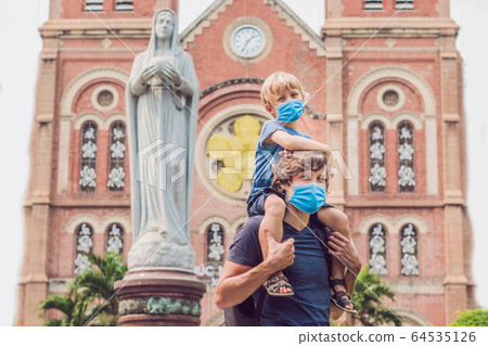 Father and son in medical mask in the background Notre dame de Saigon Cathedral, build in 1883 in Ho 64535126